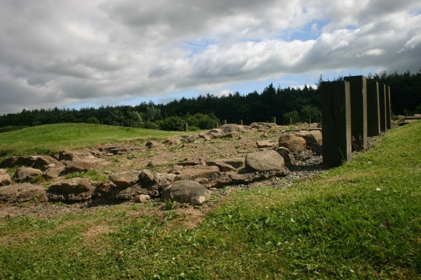 Rubble from a Roman road leading into the site of a Roman fortlet at Kinneil, Bo'ness. On the right are posts to mark buildings. In the background is woodland.